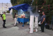 Hombre mostrando un recipiente metálico con castañas asadas junto a un puesto de venta bajo una estructura azul.